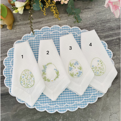Quatro guardanapos brancos bordados com flores, numerados 1 a 4, sobre tabuleiro azul e branco xadrez em superfície cinzenta