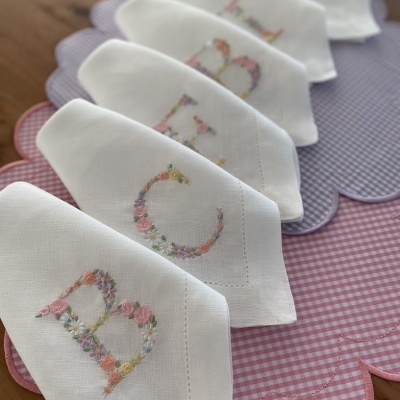 Seis guardanapos brancos bordados com letras florais sobre pano de mesa rosa xadrez e pano lilás