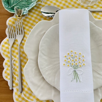 Conjunto de jantar com pratos brancos ondulados, guardanapo bordado, talheres prateados, prato verde em forma de folha e toalha de mesa xadrez amarela