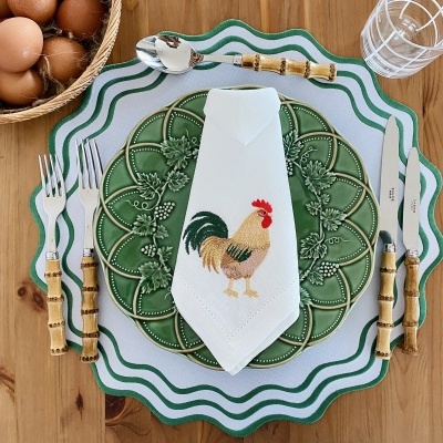 Conjunto de loiça com pratos verdes decorados, talheres com cabo de bambu, guardanapo branco com bordado de galo e copo transparente sobre mesa de madeira