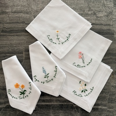 Guardanapos brancos com bordados de flores coloridas e frases em verde sobre superfície cinza.