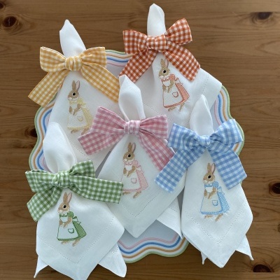Guardanapos de tecido branco com bordado de coelho e laços xadrez coloridos sobre prato decorativo