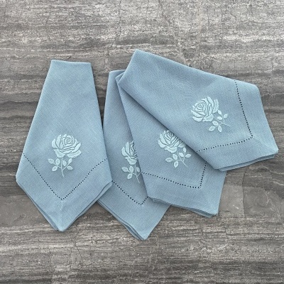 Quatro guardanapos azuis claros com rosa bordada em branco sobre superfície cinza