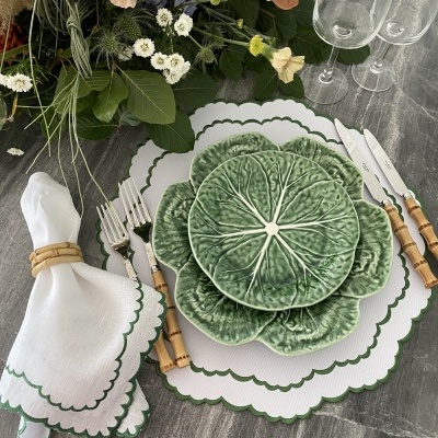 Lugar de mesa com louça verde com relevo de folhas, guardanapo branco, talheres com cabo castanho e copos de vidro numa mesa cinzenta com flores.