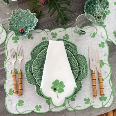 Mesa posta com loiça verde formato de folhas, guardanapo branco com bordado de trevo e joaninha, talheres com cabo de madeira e copo transparente sobre toalha bordada trevos verdes