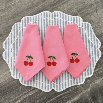 Três guardanapos cor-de-rosa com bordado de cerejas sobre suportes de tecido riscado azul e branco em superfície de pedra cinzenta.