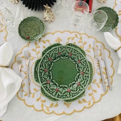 Conjunto de pratos verdes com padrão de folhas e bagas vermelhas numa mesa com toalha branca bordada a amarelo