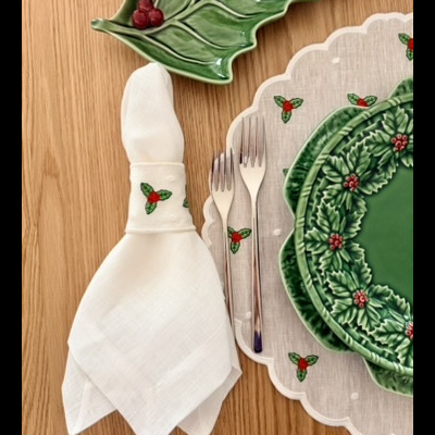 Mesa posta com pratos cerâmicos verdes com padrão de folhas, guardanapo branco com argola decorativa, talheres e toalha com estampas natalícias.