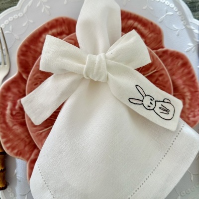 Guardanapo branco com bordado de coelho num prato vermelho e um prato branco por baixo, sobre mesa de madeira