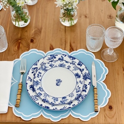 Mesa posta com pratos brancos e azuis, talheres de cabo de madeira, copos de vidro texturizados, guardanapo branco, base azul clara e flores em vasos pequenos.