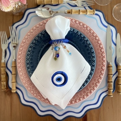 Mesa posta com pratos decorativos azul e rosa, talheres com cabo de bambu, guardanapo branco com olho turco azul e flores cor-de-rosa.