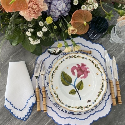 Mesa posta com loiça floral, talheres de cabo de madeira, guardanapo bordado azul e flores mistas