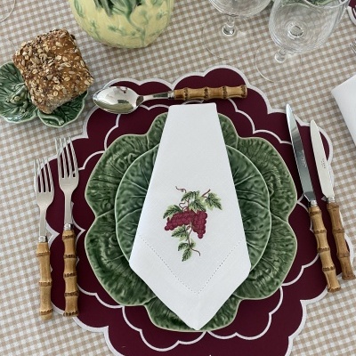 Mesa posta com pratos verdes em forma de folha, talheres com cabo tipo bambu, guardanapo branco com bordado de uvas e pão integral num pires verde