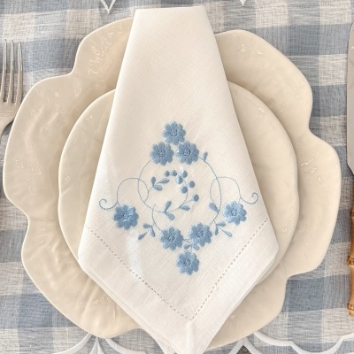 Guardanapo branco bordado com flores azuis sobre pratos brancos em toalha xadrez azul e branca.