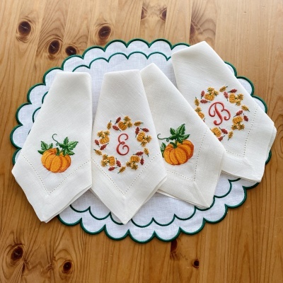 Guardanapos brancos bordados com abóboras laranja e monogramas vermelhos, em individual branco com borda verde, sobre mesa de madeira