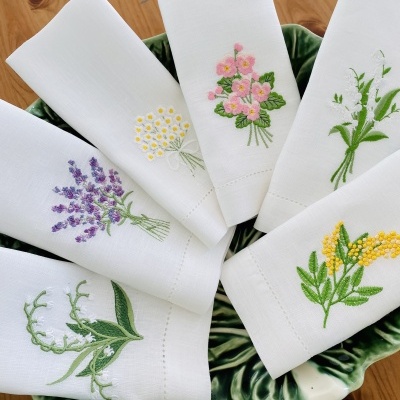 Seis guardanapos brancos bordados com flores coloridas numa travessa verde em mesa de madeira