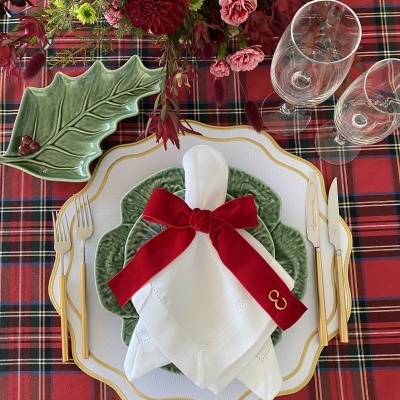 Mesa posta com pratos brancos e verdes, talheres dourados e arranjo floral sobre toalha xadrez vermelha