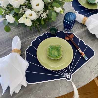 Mesa posta de jantar com prato verde, passadeira azul em forma de leque, copo azul e flores brancas