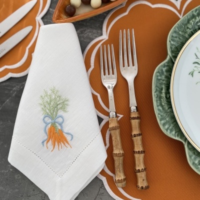 Guardanapo branco bordado com cenouras, dois garfos com cabo de madeira, individuais laranja com borda branca e prato verde sobre mesa cinza
