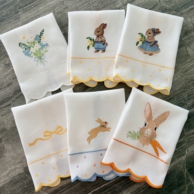 Paninhos de cozinha brancos com bordados de coelhos, flores e laços e bordas coloridas sobre superfície cinzenta.