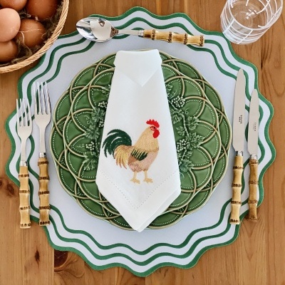 Mesa posta com pratos brancos e verdes, guardanapo bordado, talheres com cabo de bambu, ovos numa cesta e copo sobre mesa de madeira.