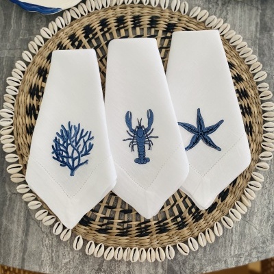 Três guardanapos brancos bordados com motivos marinhos azuis sobre prato de verga e búzios