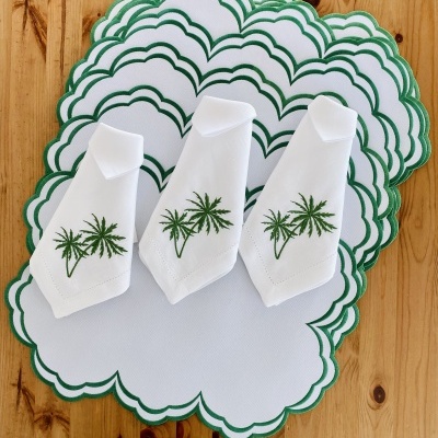 Jogo americano e guardanapos brancos com bordado de palmeira verde e bordas onduladas verdes sobre mesa de madeira.