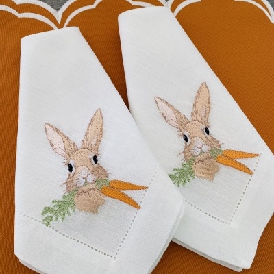 Toalhas de linho branco com bordado de coelho e cenouras, em cima de suportes castanhos com contorno branco