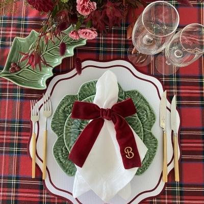 Mesa posta com conjunto de jantar em pratos brancos e verdes sobre toalha xadrez e arranjo floral vermelho