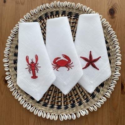 Três guardanapos brancos com bordados vermelhos de animais marinhos sobre prato trançado e conchas.