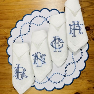 Guardanapos brancos bordados com letras azuis sobre sousplat branco com bordas azuis numa mesa de madeira