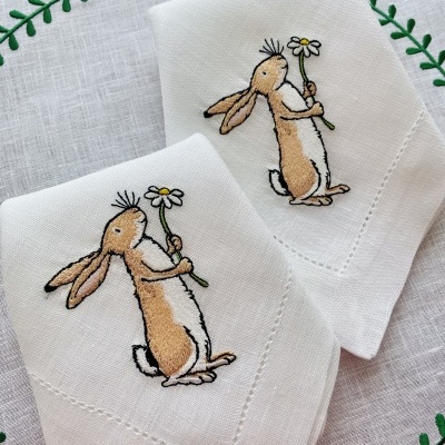 Guardanapos brancos bordados com coelhos segurando margaridas sobre tecido decorativo branco com folhas verdes.