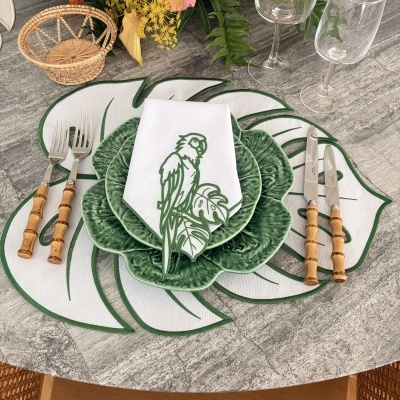 Mesa posta com prato verde em cerâmica, guardanapo branco com estampa de papagaio, talheres com cabo de madeira, copos transparentes, cesta de vime e arranjo floral
