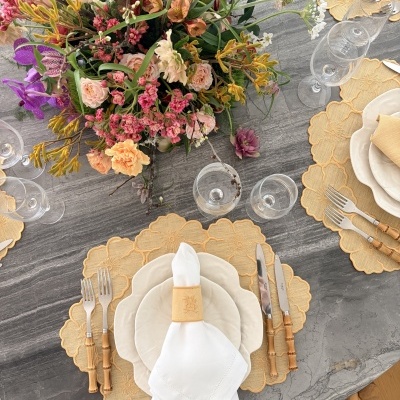 Mesa de jantar redonda em pedra cinza com louça branca, talheres com cabos em bambu, guardanapo branco, jogos americanos amarelos e arranjo de flores coloridas