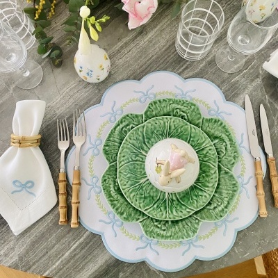 Mesa posta com pratos de cerâmica em forma de folhas verdes, talheres de cabo de madeira, guardanapo branco com bordado azul e anel dourado, copos transparentes e vaso branco com flores.
