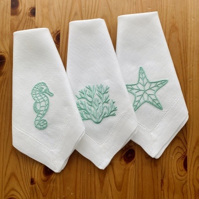 Três toalhas brancas com bordados verdes de mariscos sobre uma mesa de madeira