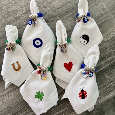 Conjunto de guardanapos brancos com bordados coloridos e anéis com pendentes sobre superfície cinza.