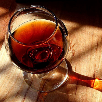 Copo de vidro com bebida castanha sobre mesa de madeira clara com sombras e reflexos