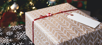 Caixa de presente bege com padrão branco, fita vermelha e etiqueta branca perto de árvore de Natal