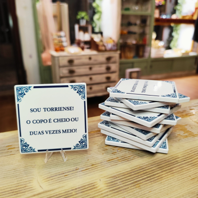 Azulejo quadrado branco com moldura azul e texto em português sobre suporte, empilhados numa mesa de madeira