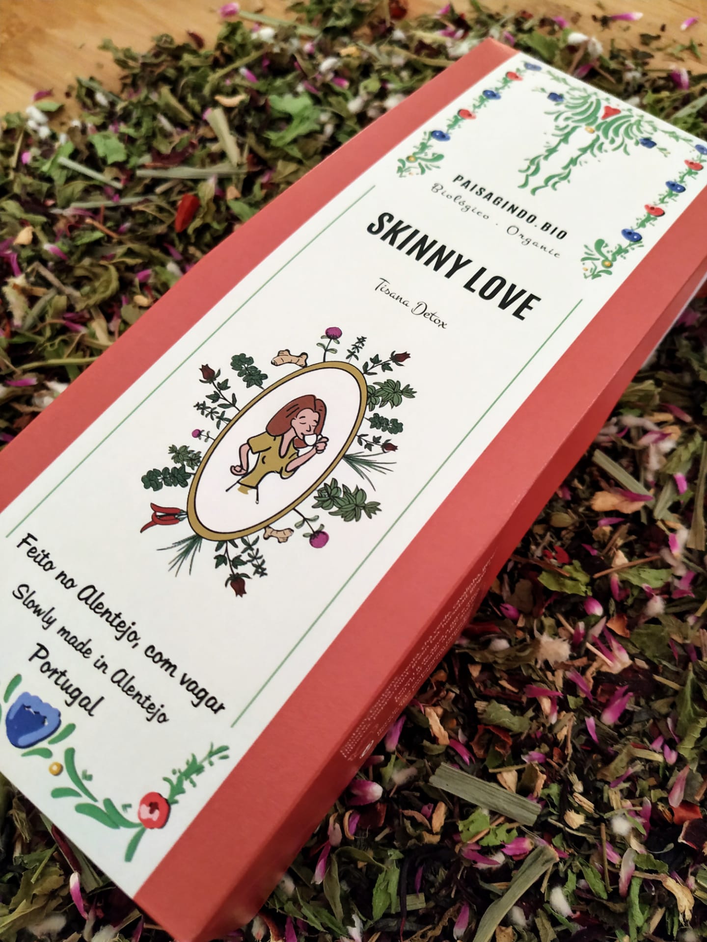 Embalagem vermelha de tisana com rótulo branco decorado e texto em português, rodeada por ervas secas e pétalas.