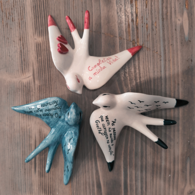 Três pássaros em cerâmica pintada com textos manuscritos, dispostos sobre madeira