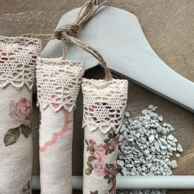 Sachês decorativos em tecido floral com renda pendurados em cabide cinza com seixos ao lado