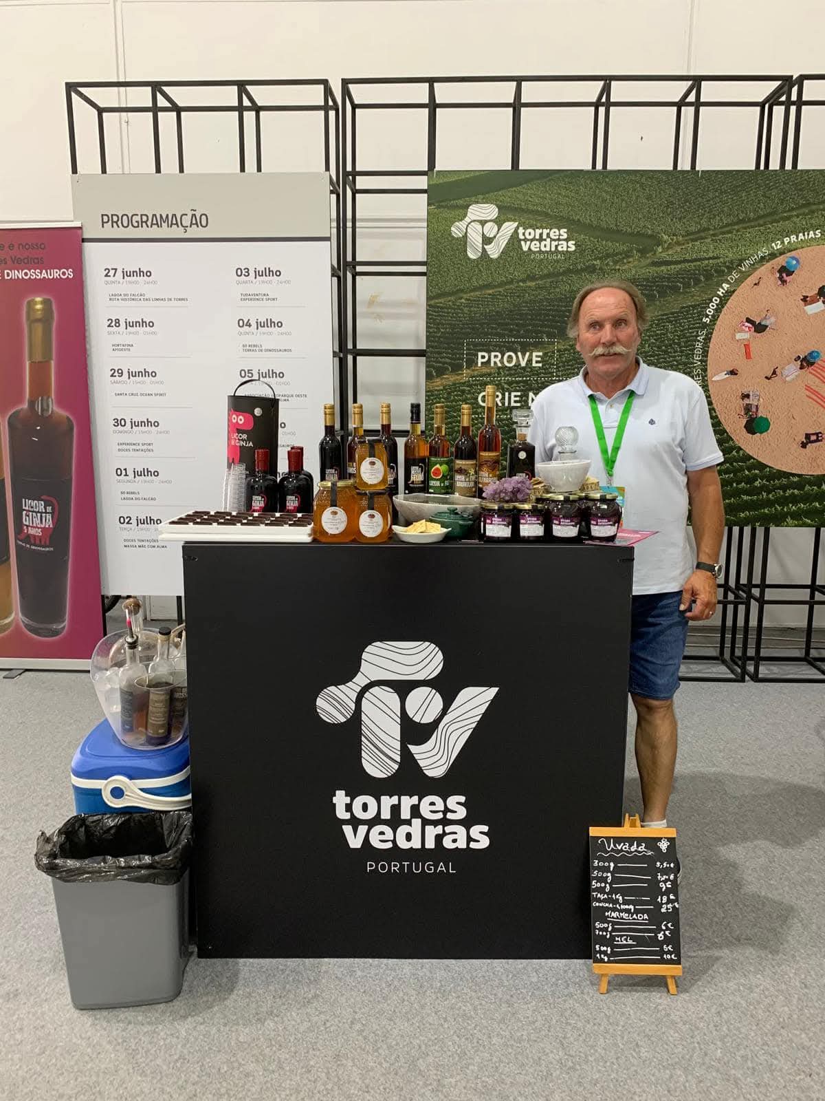 Banca preta com bebidas e compotas, homem ao lado e painéis informativos ao fundo