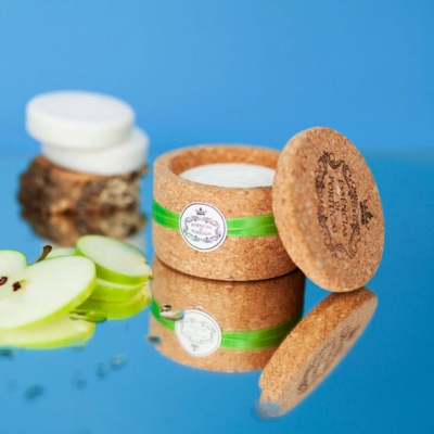 Embalagem de cortiça redonda com creme branco ao lado de fatias de maçã verde sobre fundo azul reflexivo