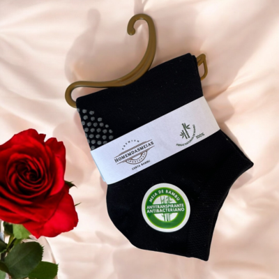 Meias pretas de bambu com etiqueta e selo verde, sobre tecido rosa claro, com rosa vermelha ao lado.