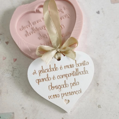 Etiqueta de coração branca com texto dourado e fita bege, sobre superfície clara com molde de silicone rosa em forma de coração ao fundo.