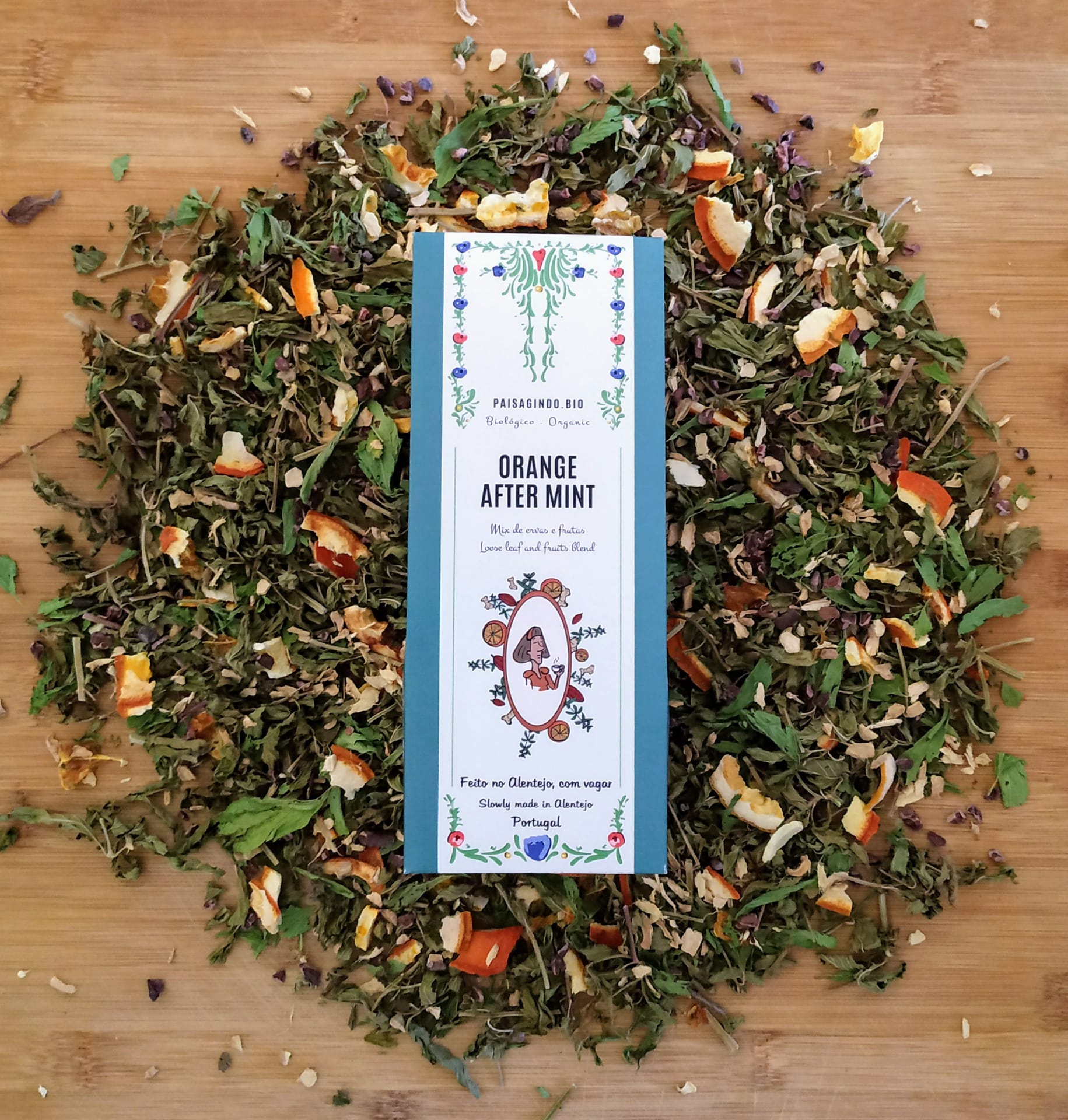 Pacote de chá Orange After Mint rodeado por ervas e cascas de laranja sobre madeira