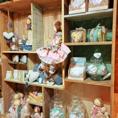 Estante de madeira com bonecas de pano, corações de tecido, frascos de vidro e caixas de sabonete artesanal