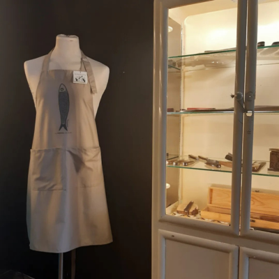 Avental bege com desenho de peixe azul em manequim ao lado de vitrine branca